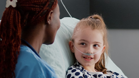 A young blonge girl sits up in a hospital bed. She has a tube providing oxygen to her now and is looking and speaking with a nurse kneeling by her bed.